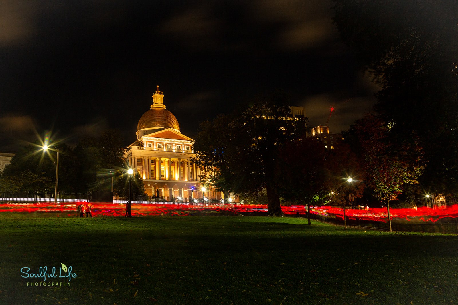 Nighttime image of Massachusetts State house lit up with warm yellow lights. Time-lapse of participants walking in front of the state house makes a red blur across the bottom of the image. 